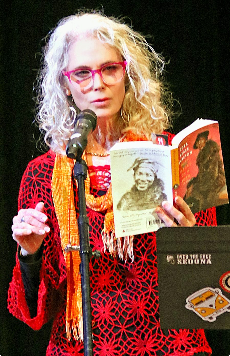 Ché reading at an Over the Edge Sedona literary event, in pink glasses and a red scarf